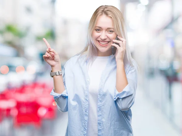 年轻的金发女人说话使用 Smarpthone 在孤立的背景非常高兴地指着手和手指的侧面 — 图库照片