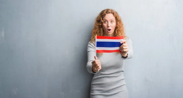 Joven Pelirroja Sobre Pared Grunge Gris Sosteniendo Bandera Tailandia Asustada —  Fotos de Stock