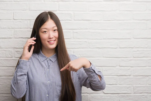 自分自身に驚き顔人差し指と電話で話しているレンガの壁を越えて若い中国の女性 — ストック写真
