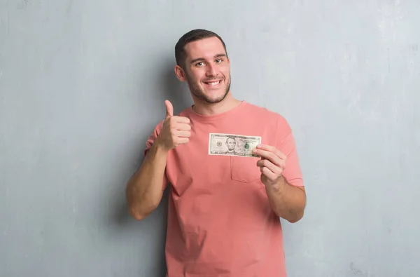 ドルの大きな満足を示す灰色グランジ壁を越えて若い白人男笑顔 の標識を行う 優秀な兆候の指で親指 — ストック写真