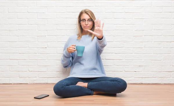 Beautiful young woman sitting on the floor driking cup of coffee at home with open hand doing stop sign with serious and confident expression, defense gesture