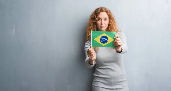 灰色垃圾墙上的年轻红头发的女人持有巴西国旗自信的表情对聪明的面孔认真思考 — 图库照片