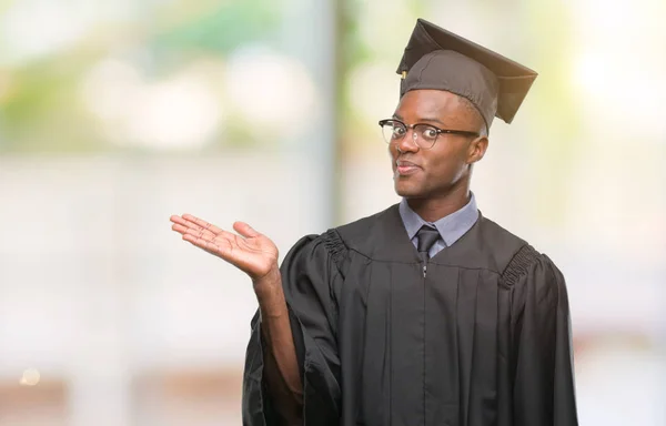 若い陽気な提示とカメラを見て手の手のひらで指している笑みを浮かべて孤立の背景の上のアフリカ系アメリカ人の卒業 — ストック写真