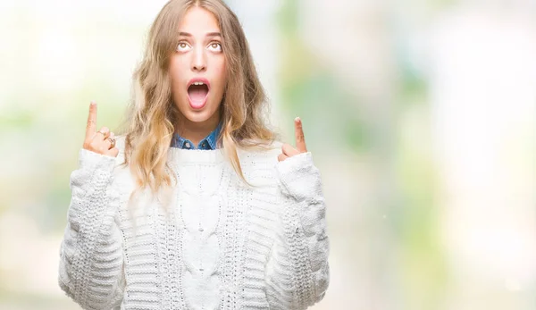 Beautiful young blonde woman wearing winter sweater over isolated background amazed and surprised looking up and pointing with fingers and raised arms.