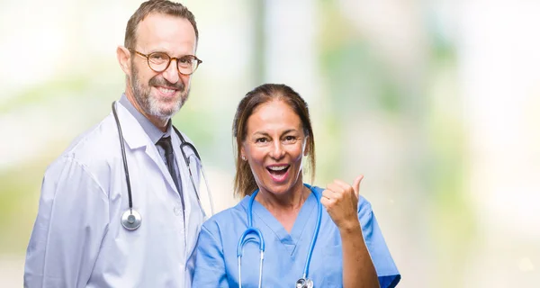 中年拉美裔医生搭档夫妇穿着医疗制服在孤立的背景微笑着快乐的脸看着 并指向侧面与拇指向上 — 图库照片
