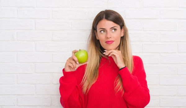 年轻的成年妇女在白色砖墙吃新鲜的绿色苹果严肃的面孔思考问题 非常混淆的想法 — 图库照片