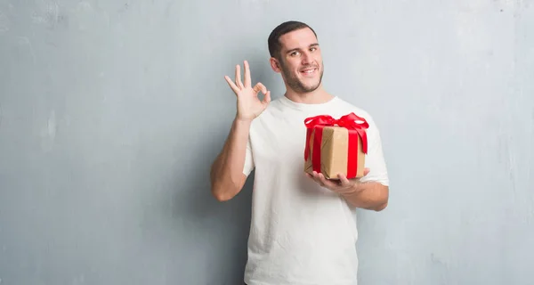 プレゼントを保持している灰色のグランジの壁を越えて若い白人男優秀なシンボルの指で サインをしています — ストック写真