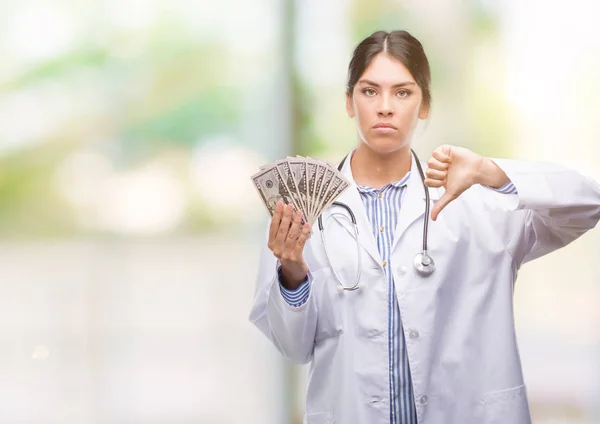 Young hispanic doctor woman holding dollars with angry face, negative sign showing dislike with thumbs down, rejection concept