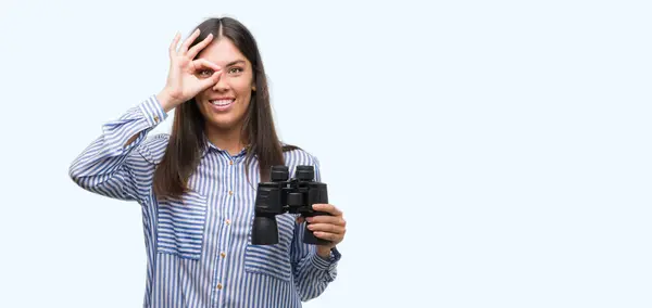 年轻的西班牙裔妇女拿着双筒望远镜快乐的脸微笑着做 签名用手在眼睛看通过手指 — 图库照片