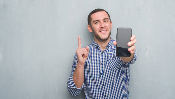 Молодой кавказский мужчина над серой гранж-стеной показывает чистый экран смартфона удивленный идеей или вопросом указывая пальцем со счастливым лицом, номер один
