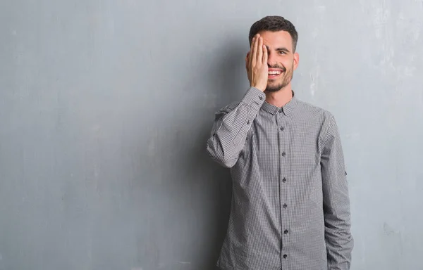 年轻的男子站在灰色的垃圾墙覆盖一只眼睛与手自信的脸上的笑容和惊喜的情绪 — 图库照片