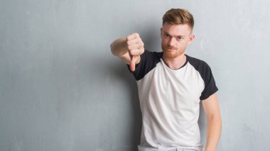 Genç Kızıl saçlı adam mutsuz ve kızgın ret gösterilen ve başparmak aşağı hareketi ile negatif seyir gri grunge duvarın üzerinden. Kötü ifade.