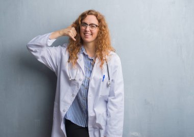 Genç redhead doktor kadın yapıyor gülümseyen bir palto giyiyor gri grunge duvarın üzerinden telefon hareketi ile el ve parmak telefonda konuşuyor gibi. Kavramları iletişim.