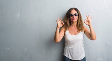 Orta yaş İspanyol kadın güneş gözlüğü takıyor gri duvarın üzerinden rahatla ve gülümseyen gözlerle kapalı parmak jestle meditasyon yapıyor. Yoga kavramı.