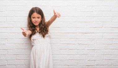 Genç İspanyol çocuğun el ile pozitif jest yapıyor onaylama beyaz tuğla duvarın üzerinden başparmak gülümseyen ve başarı için mutlu. Kamera, kazanan jest arıyorsunuz.