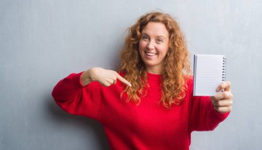Gri grunge duvar gösteren boş defter kendisi için sürpriz yüz işaret parmağı ile genç Kızıl saçlı kadın
