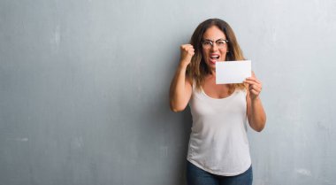 Üzerinde gri grunge duran orta yaş İspanyol kadın duvar boş kartı rahatsız tutarak ve boşa çıkarmak ile öfke, çılgın ve yükseltilmiş eliyle diye bağıran öfke kavramı bağırarak.