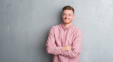 Gri grunge genç Kızıl saçlı adam kameraya bakarak çapraz silah ile gülümseyen pembe gömlek giyen mutlu yüz duvar. Olumlu bir kişi.