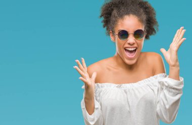 Genç afro Amerikalı kadın deli ve silah ile başarı için deli kutluyor izole arka plan üzerinde güneş gözlüğü takıyor kaldırdı ve heyecanlı çığlık gözler kapalı. Kazanan kavramı