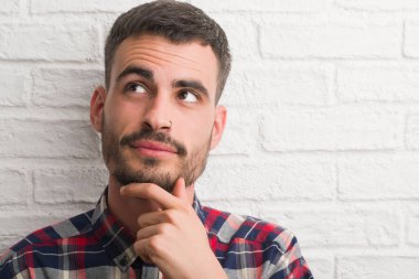 Genç yetişkin adam beyaz tuğla duvar ciddi bir yüz düşünme hakkında soru, çok karışık bir fikir üzerinde ayakta