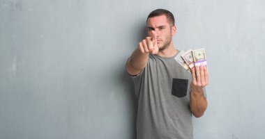 Beyaz delikanlı paralarını kamera ve size parmak ile işaret tutan gri grunge duvarın üzerinden, el işareti, pozitif ve kendinden emin bir jest ön