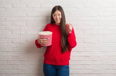 Genç Çinli kadın ayakta ve diş gösterilen kendine güvenen bir gülümsemeyle gülümseyerek mutlu bir yüz ile pop Mısır yeme tuğla duvarın üzerinden