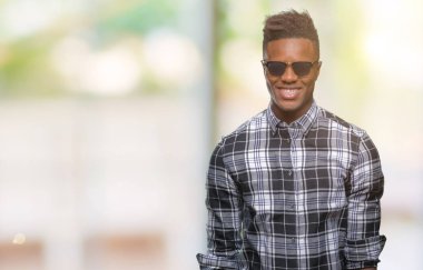 Genç Afro-Amerikan adam yüzünde mutlu ve güzel bir gülümseme ile izole arka plan üzerinde güneş gözlüğü takıyor. Şanslı kişi.