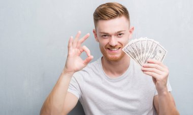 Genç Kızıl saçlı adam parmak, mükemmel sembolü ile ok işareti yapıyor dolar tutan gri grunge duvarın üzerinden