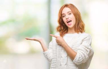 İzole arka plan giyen kış kazak hayret ve el ile sunulması ve parmak ile işaret ederken kameraya gülümseyen genç güzel kadın.