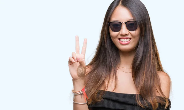若いアジア女性の勝利のサインを行うカメラでウィンクしている幸せそうな顔を浮かべて孤立の背景の上にサングラスをかけています — ストック写真