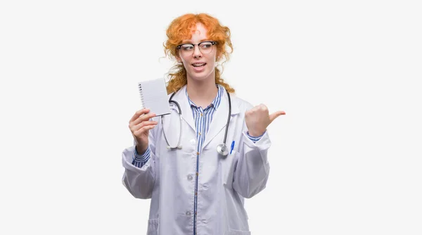 指していると幸せそうな顔を笑顔で側まで親指で示す空白のノートブックを保持している赤毛の若い医師女性 — ストック写真