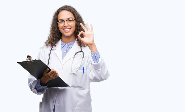 年轻的西班牙裔医生的女人拿着剪贴板做 标志与手指 优秀的符号 — 图库照片