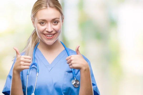 Young blonde surgeon doctor woman over isolated background success sign doing positive gesture with hand, thumbs up smiling and happy. Looking at the camera with cheerful expression, winner gesture.