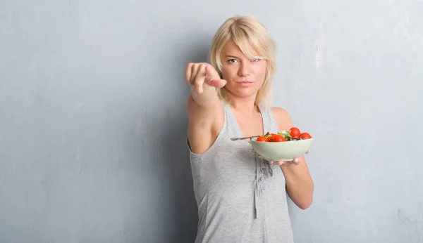 成年白种人妇女在垃圾灰墙吃西红柿沙拉指向相机和你 正面和自信的手势从前面 — 图库照片