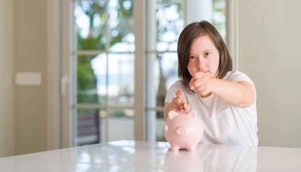 Down syndrome woman at home holding piggy bank pointing with finger to the camera and to you, hand sign, positive and confident gesture from the front