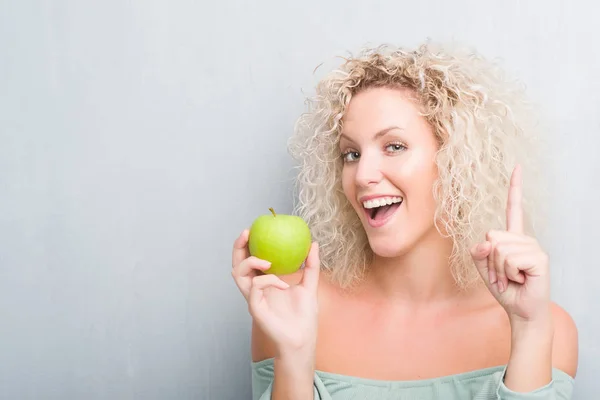 年轻的金发女子超过难看的东西灰色背景吃绿色苹果惊讶与一个想法或问题指向手指与幸福的脸 — 图库照片