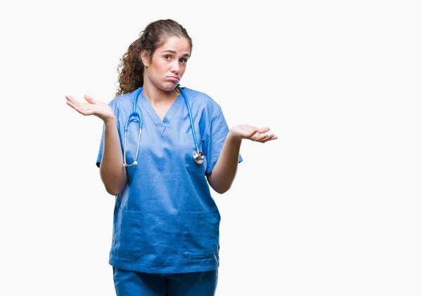 年轻的黑发医生女孩穿着护士或外科医生制服在孤立的背景无线索和混乱的表情与手臂和手抬起来 怀疑概念 — 图库照片