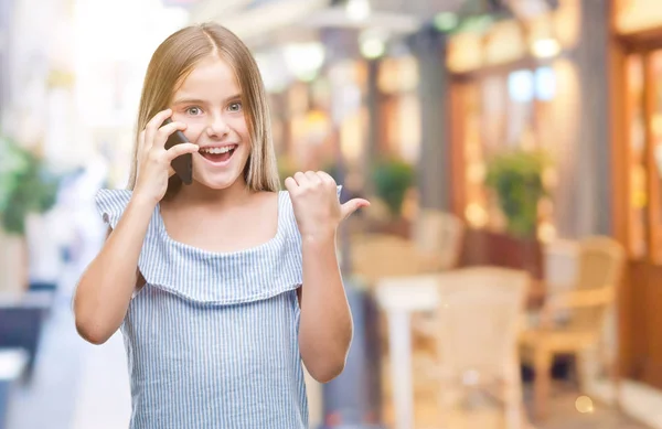 年轻美丽的女孩在电话上谈论孤立的背景指向和显示与拇指到一边与愉快的脸微笑 — 图库照片