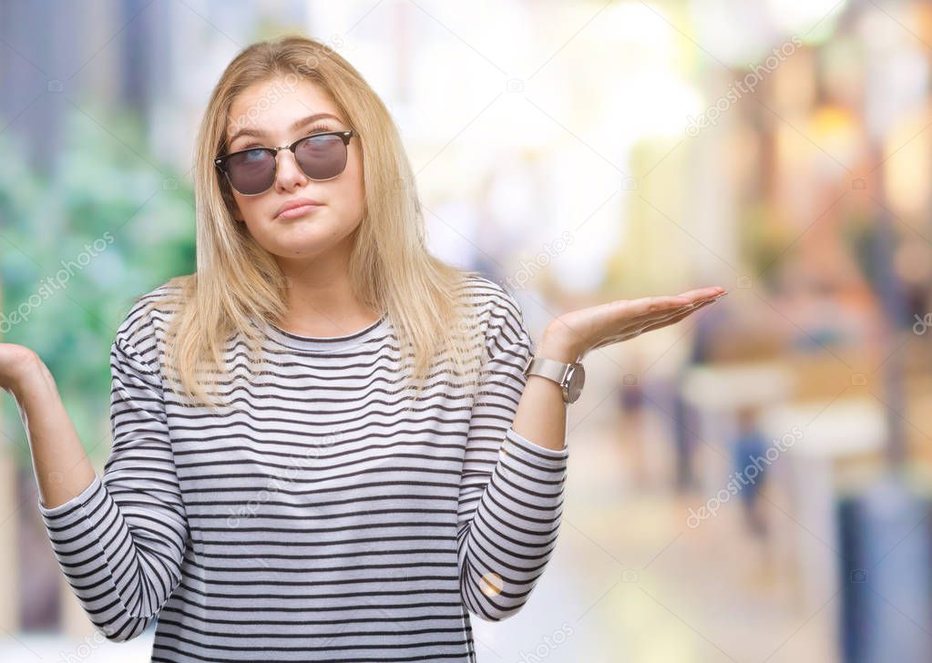 Young caucasian woman wearing sunglasses over isolated background clueless and confused expression with arms and hands raised. Doubt concept.