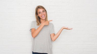 Güzel genç kadın hayrete ve el ile sunulması ve parmak ile işaret ederken kameraya gülümseyen beyaz tuğla duvarın üzerinden.