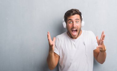 Yakışıklı genç gri grunge üzerinde duvar giyen kulaklık büyük gülümseme ile çığlık zaferini kutluyor ve ellerini kaldırdı müzik çok mutlu ve heyecanlı, kazanan ifade dinleme