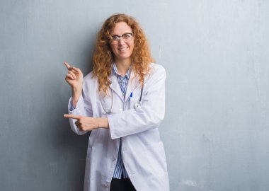 Genç redhead doktor kadın gülümseyerek ve iki elleri ve parmakları ile tarafına işaret kamera bakarak bir palto giyiyor gri grunge duvarın üzerinden.