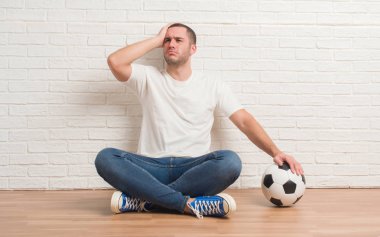 Futbol futbol topu kafa üzerinde el ile vurguladı holding beyaz tuğla duvar üzerine oturan genç beyaz adam şok utanç ve sürpriz suratla, öfkeli ve sinirli. Korku ve hata için üzgün.