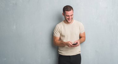 Gri grunge üzerinde beyaz delikanlı duvar manifatura ayakta ve diş gösterilen kendine güvenen bir gülümsemeyle gülümseyerek mutlu bir yüz ile smartphone kullanarak bir ileti