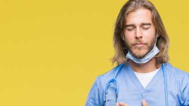 Göğüs kapalı gözlerle ellerinde ve yüzünde minnettar jest ile gülümseyen izole arka plan üzerinde uzun saçlı Genç yakışıklı Doktor adam. Sağlık kavramı.