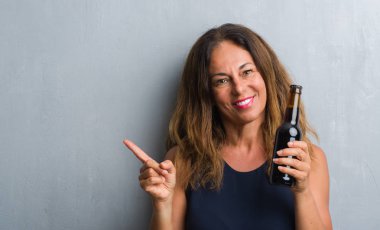 Bira şişesi çok mutlu el ve parmak ile tarafına işaret tutan gri grunge duvarın üzerinden duran orta yaş İspanyol kadın