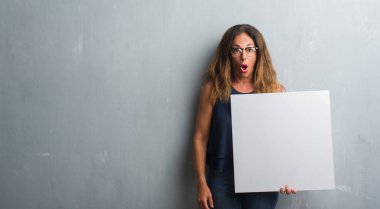 Gri grunge duvar holding boş afiş bir sürpriz yüz, korkuyor ve heyecanlı korku ifadesi ile şok korkmuş üzerinde duran orta yaş İspanyol kadın