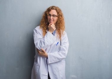 Genç redhead doktor kadın gri grunge duvar emin çapraz kollar ve çeneme kaldırdı el ile gülümseme ile kameraya bakarak bir palto giyiyor. Pozitif düşünce.