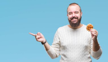 Genç beyaz hipster man izole arka tarafına el ve parmak ile işaret çok mutlu üzerinde tatlı waffle yemek