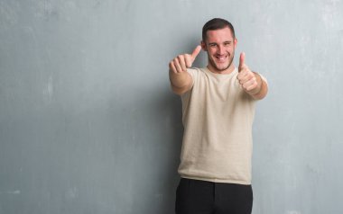 Gri grunge duvar onaylama üzerinde beyaz delikanlı el ile pozitif jest yapıyor gülümseyen ve başarı için mutlu yaşasın. Kamera, kazanan jest arıyorsunuz.